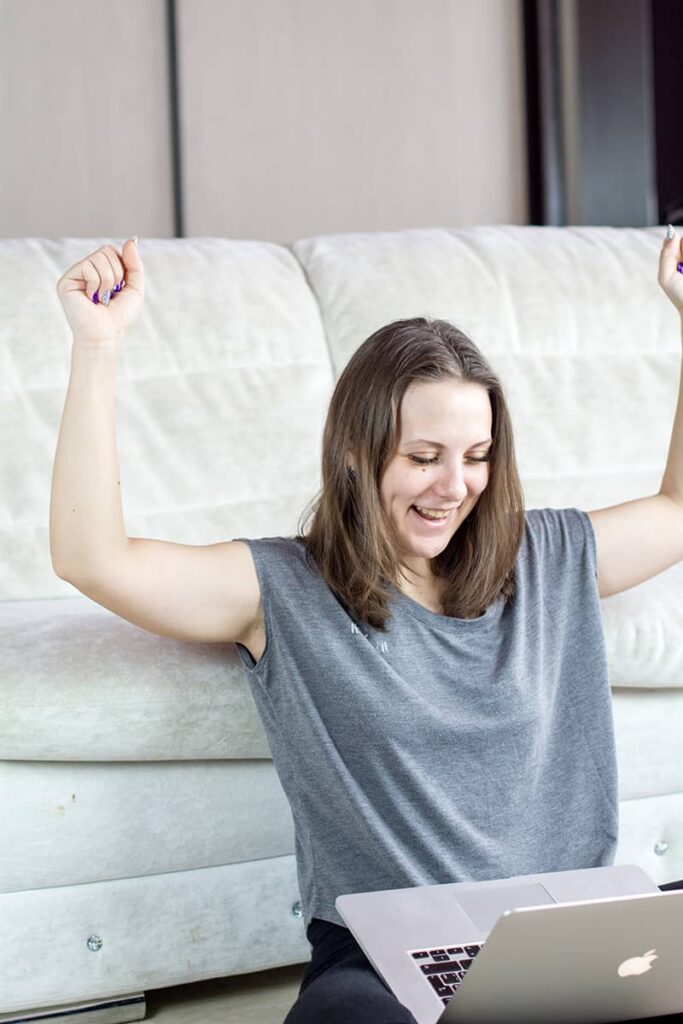 A girl sitting on the floor with her laptop and arms in the air celebrating